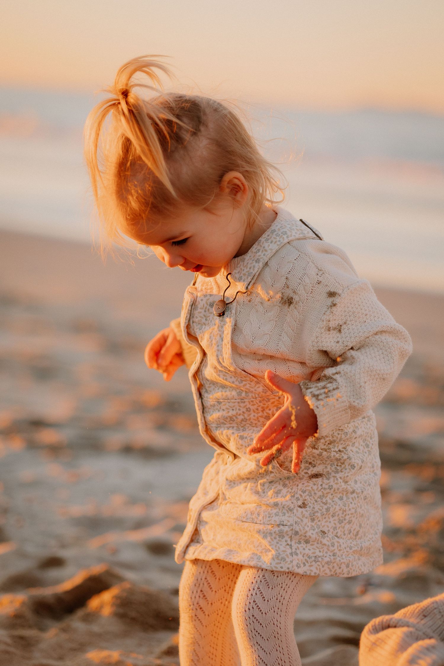 A tender beach moment at sunset with a toddler and adult sitting together on golden sand by the ocean.