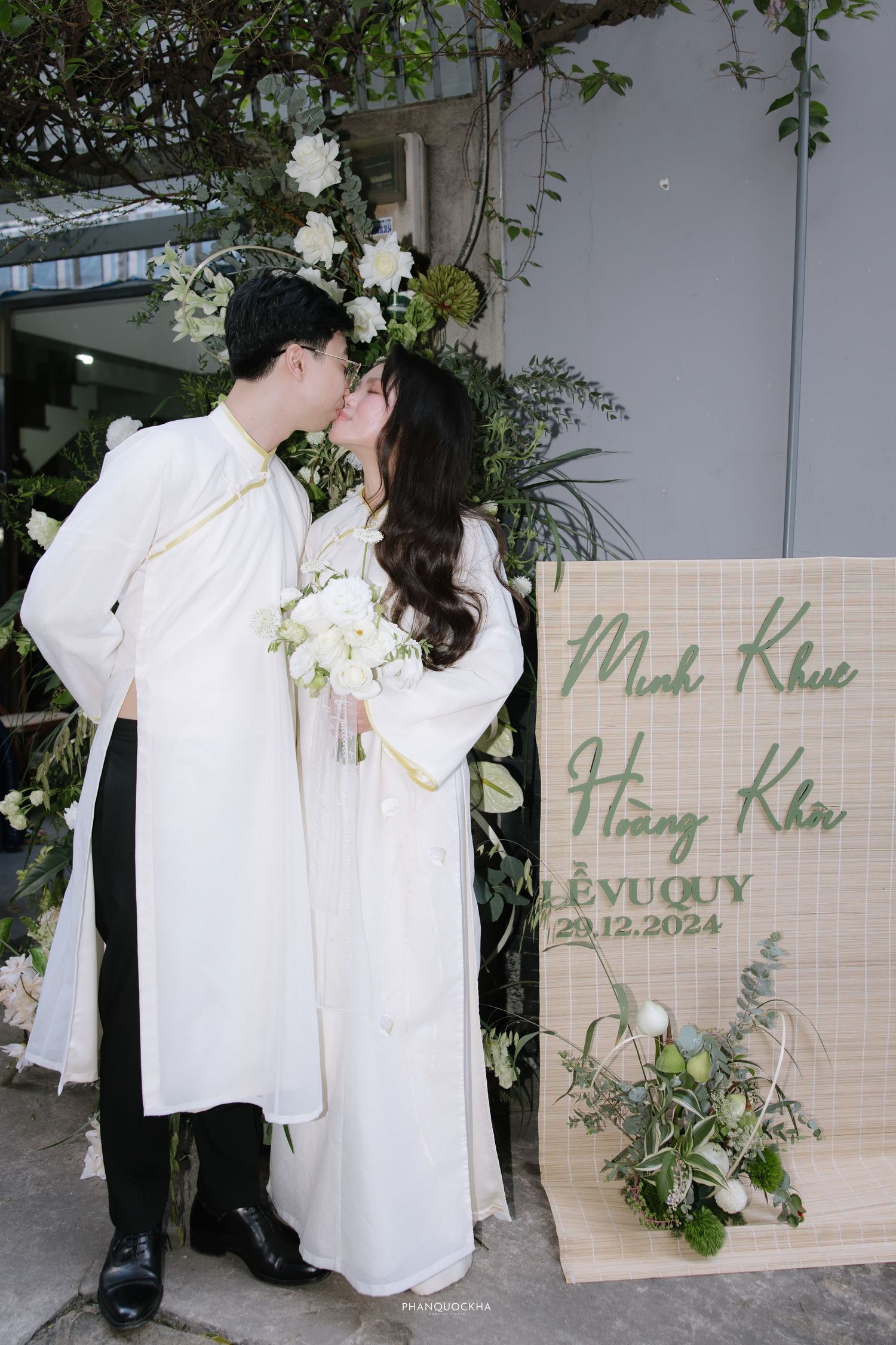 Vietnamese couple shares a romantic kiss beside their wedding sign surrounded by white floral arrangements.