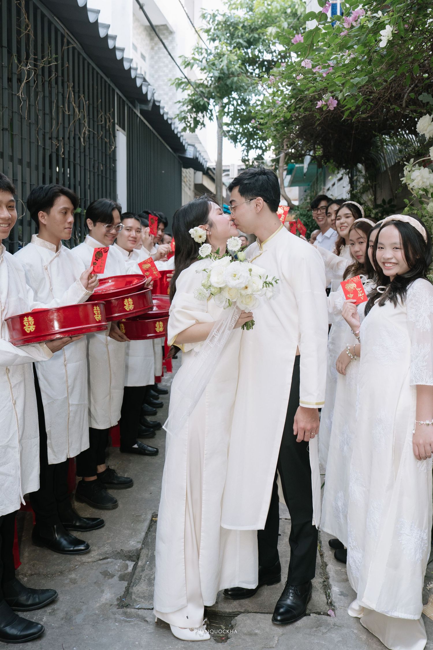 Vietnamese bride and groom share a kiss surrounded by wedding party holding red envelopes and gift trays.