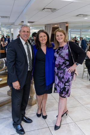Three people in formal attire standing together at an event in a room with tiled floors.
