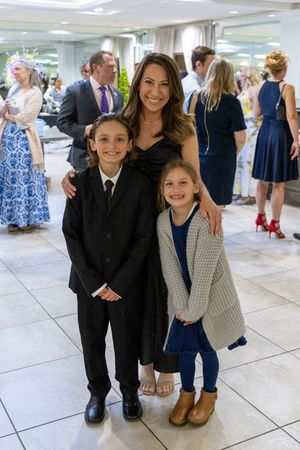 Children in formal wear gather together at an event on white tiled floors in front of glass walls.