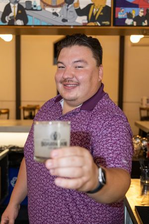 Person in purple patterned polo shirt holds up a glass while smiling at a bar setting.