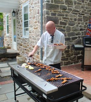 A row of grills with food being cooked on a stone patio next to a rustic building.