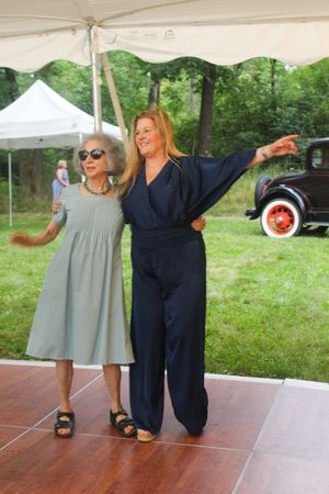 Two women strike playful poses on a dance floor with vintage car visible in background.