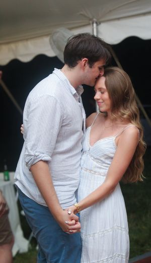 A romantic couple shares a tender moment under a white tent at an outdoor celebration.