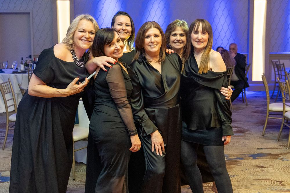 Group of six women in black dresses smiling and posing together at a formal event in a ballroom.