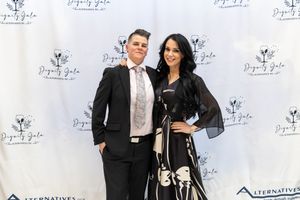 A couple poses together at a red carpet event with repeating logo backdrop.