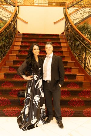 Elegant couple posing in formal attire on grand staircase with ornate iron railings and red floral carpet.
