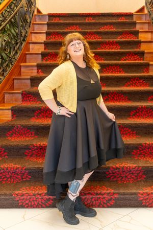 Woman in black dress and yellow cardigan poses on ornate staircase with red floral carpet.