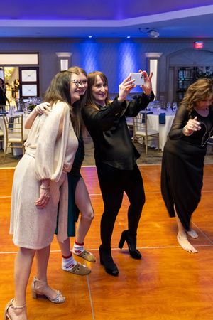 People taking selfies and dancing on a wooden dance floor at an event venue with blue lighting and table settings.