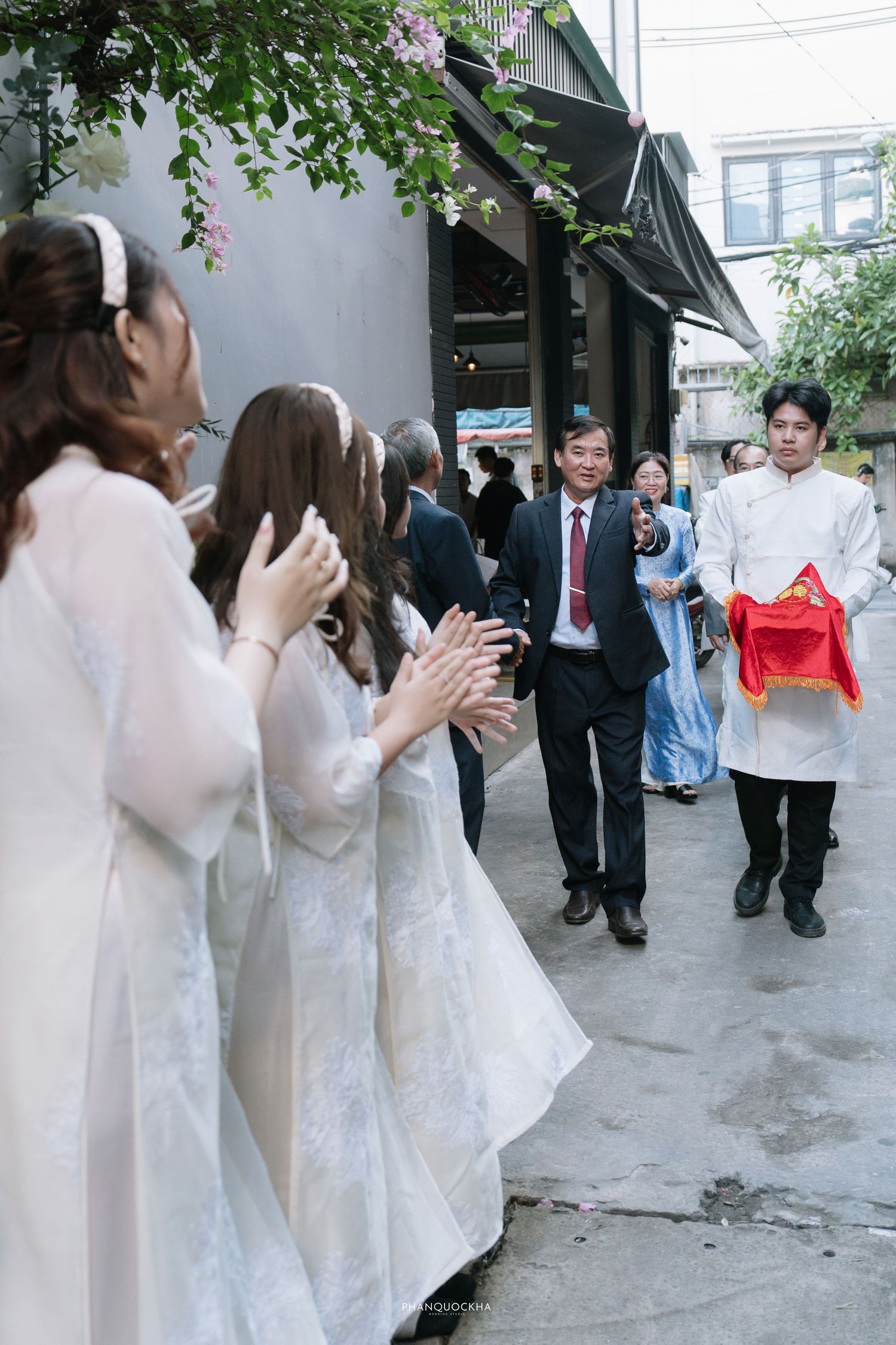 Wedding procession arrives as women in white ao dai clap in welcome outside decorated entrance.