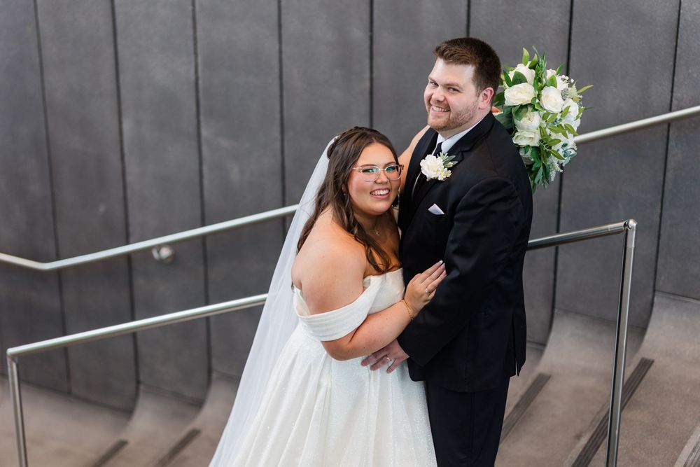 A happy couple embraces on concrete stairs during their wedding day, with white floral bouquet and classic attire.