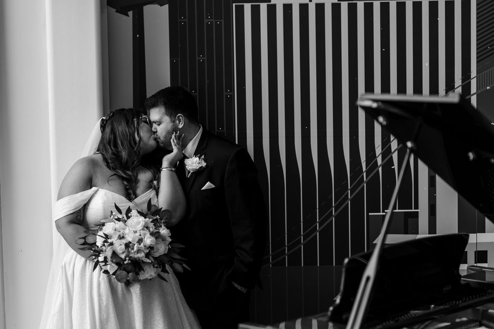 Black and white wedding photograph showing a couple embracing in a romantic moment with vertical window blinds.
