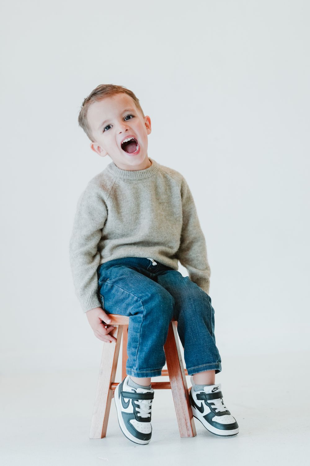 Toddler in grey sweater and jeans sitting on wooden stool against white background.