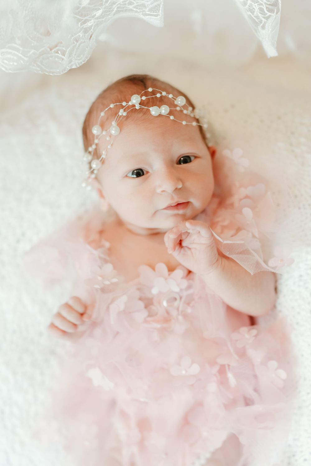 Baby wearing delicate white lace dress with pearl headpiece on white background.