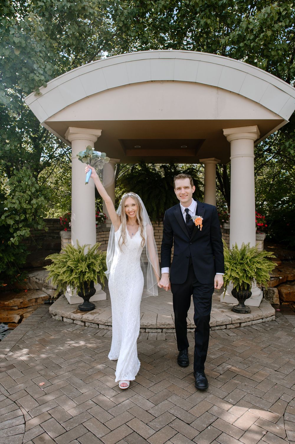 A wedding couple walks under an elegant outdoor stone gazebo with white columns and lush greenery.