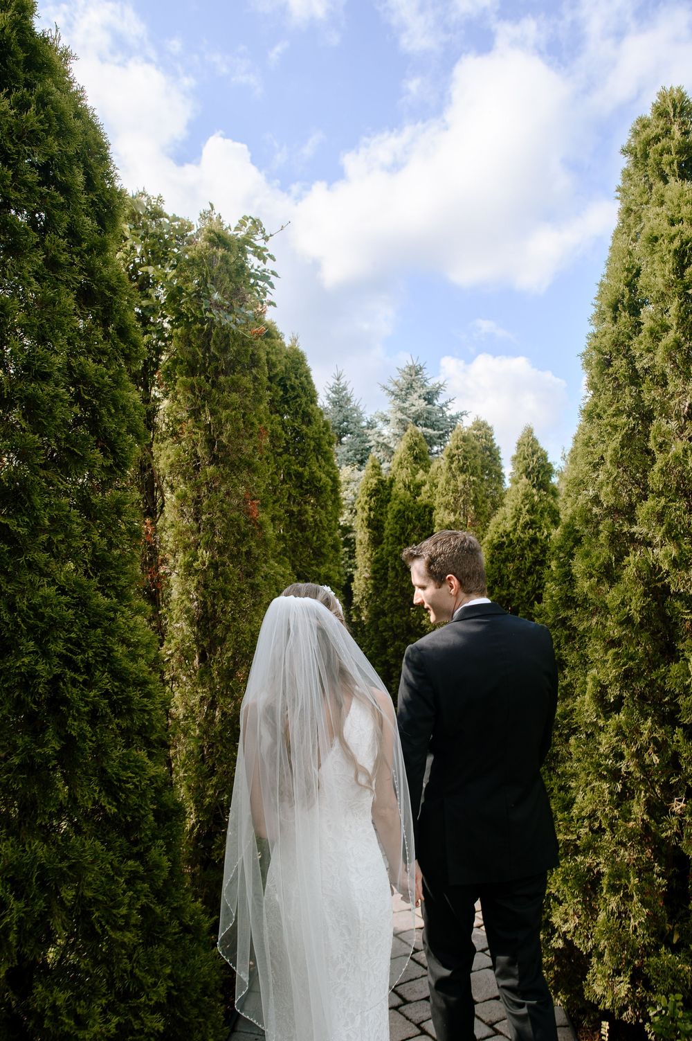 A bride and groom walk through a path lined with tall evergreen trees on their wedding day.