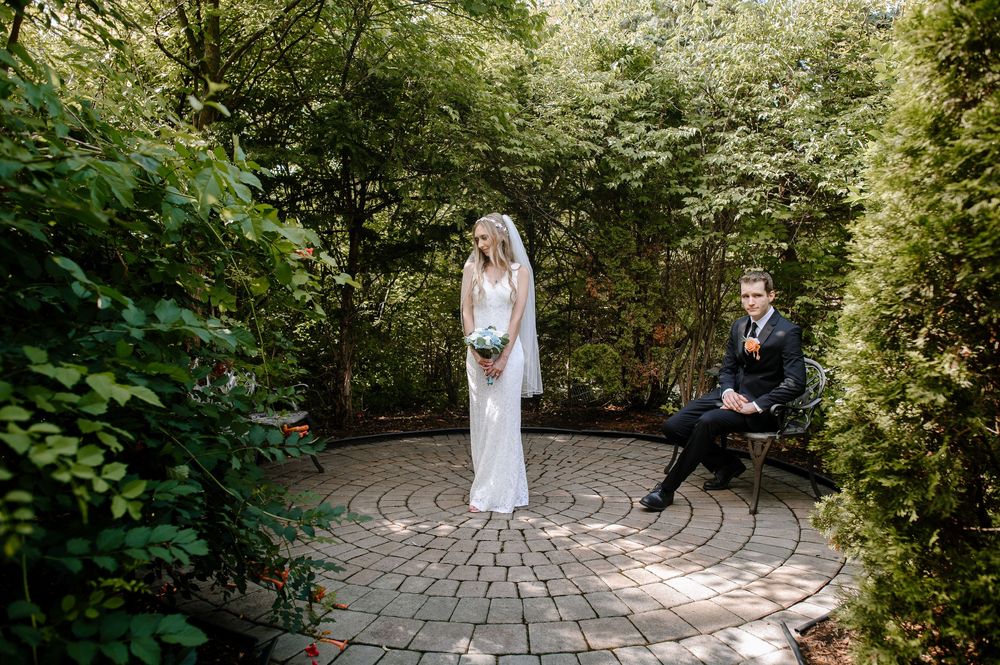 Couple in wedding attire poses in a romantic garden setting with circular brick patio surrounded by greenery.