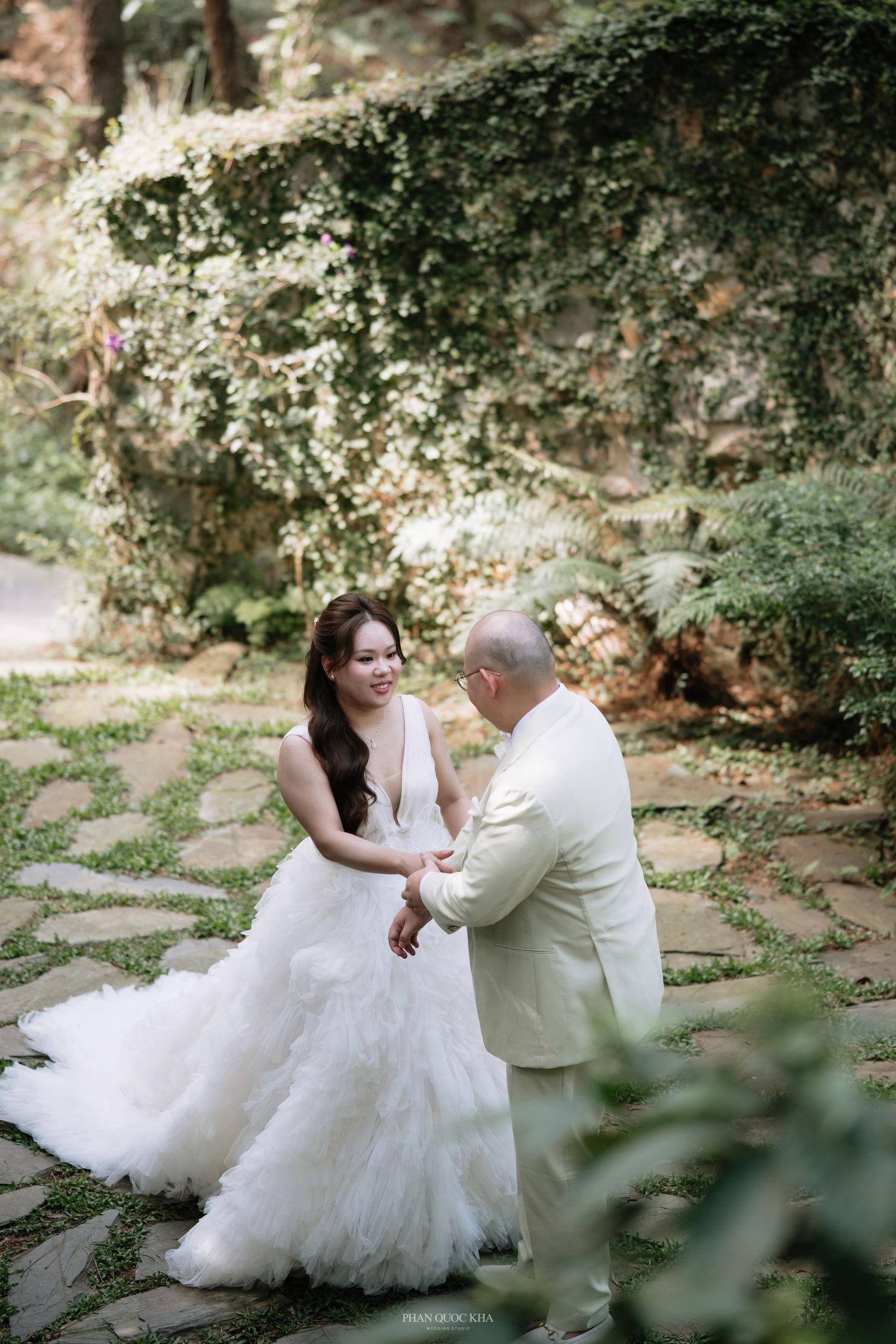 Bride in a deep-V ruffled gown holds hands with groom in cream suit in ivy-covered garden.