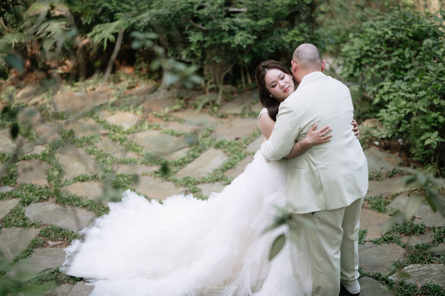 Bride in a flowing white gown embracing groom in a light suit in a garden setting.