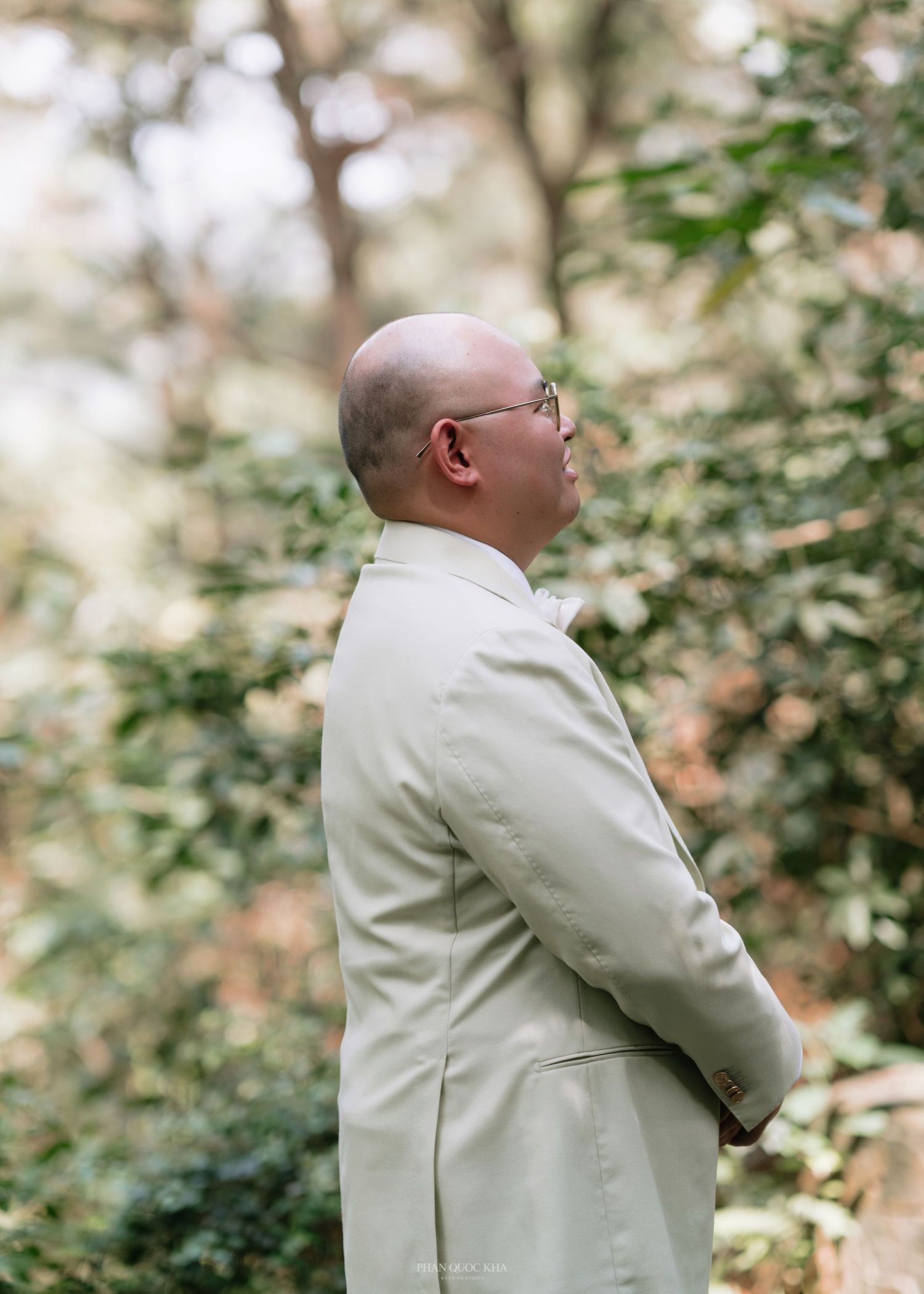 Groom in sage green suit standing in profile amid lush garden greenery before wedding ceremony.