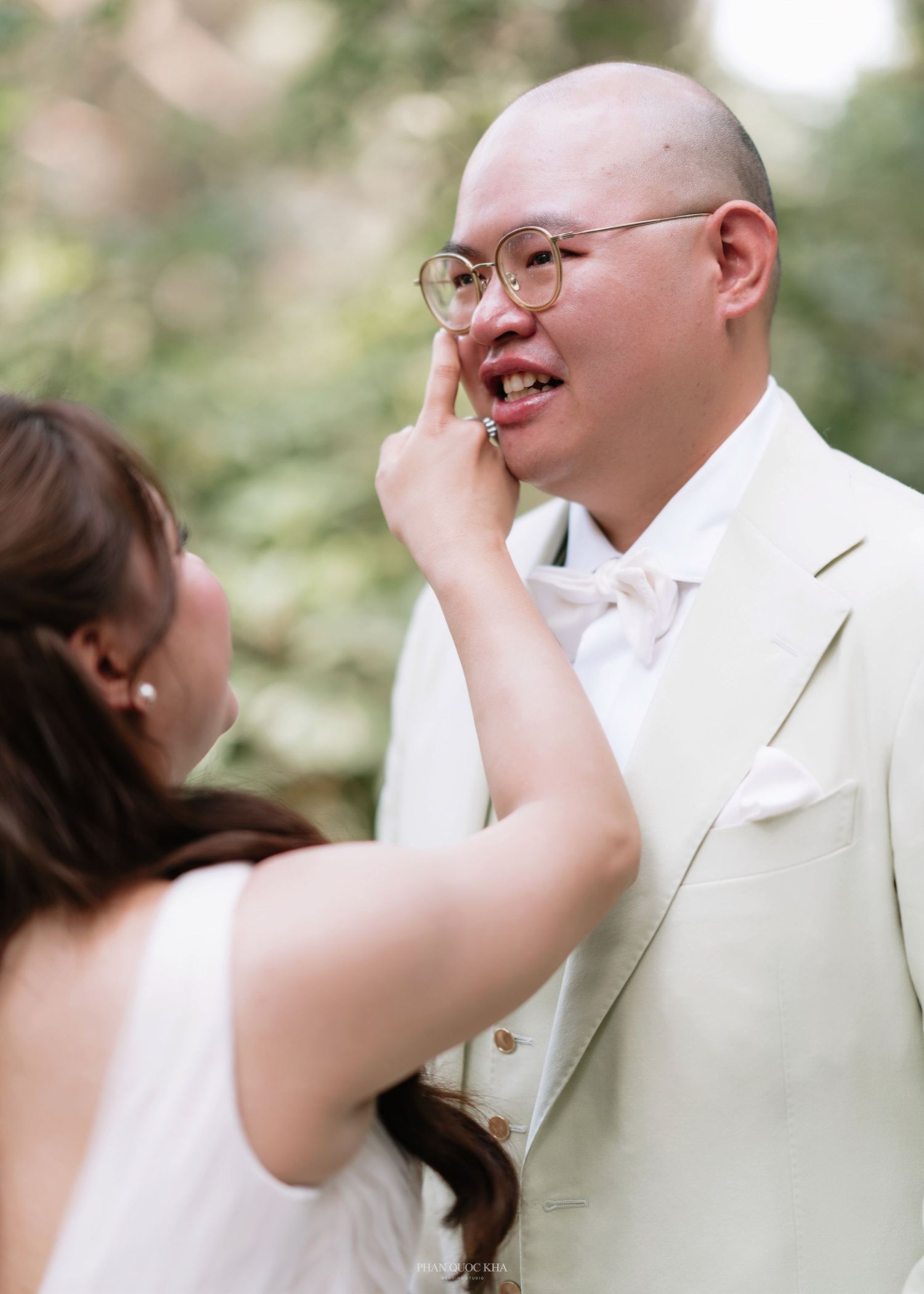 Groom in cream suit smiling emotionally as bride wipes tear during outdoor wedding first look.