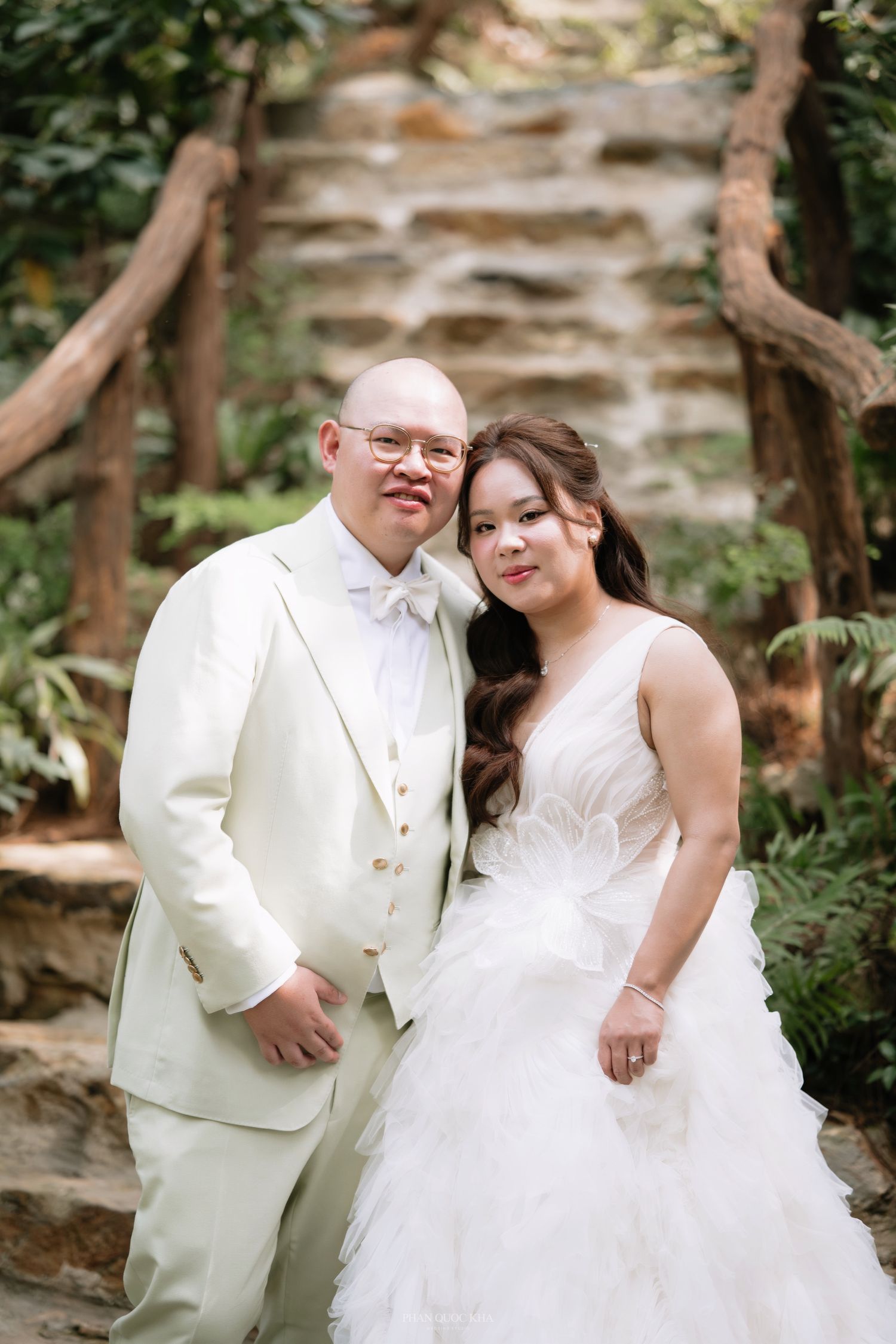 Smiling Asian bride and groom in cream wedding attire posing together in front of stone waterfall.