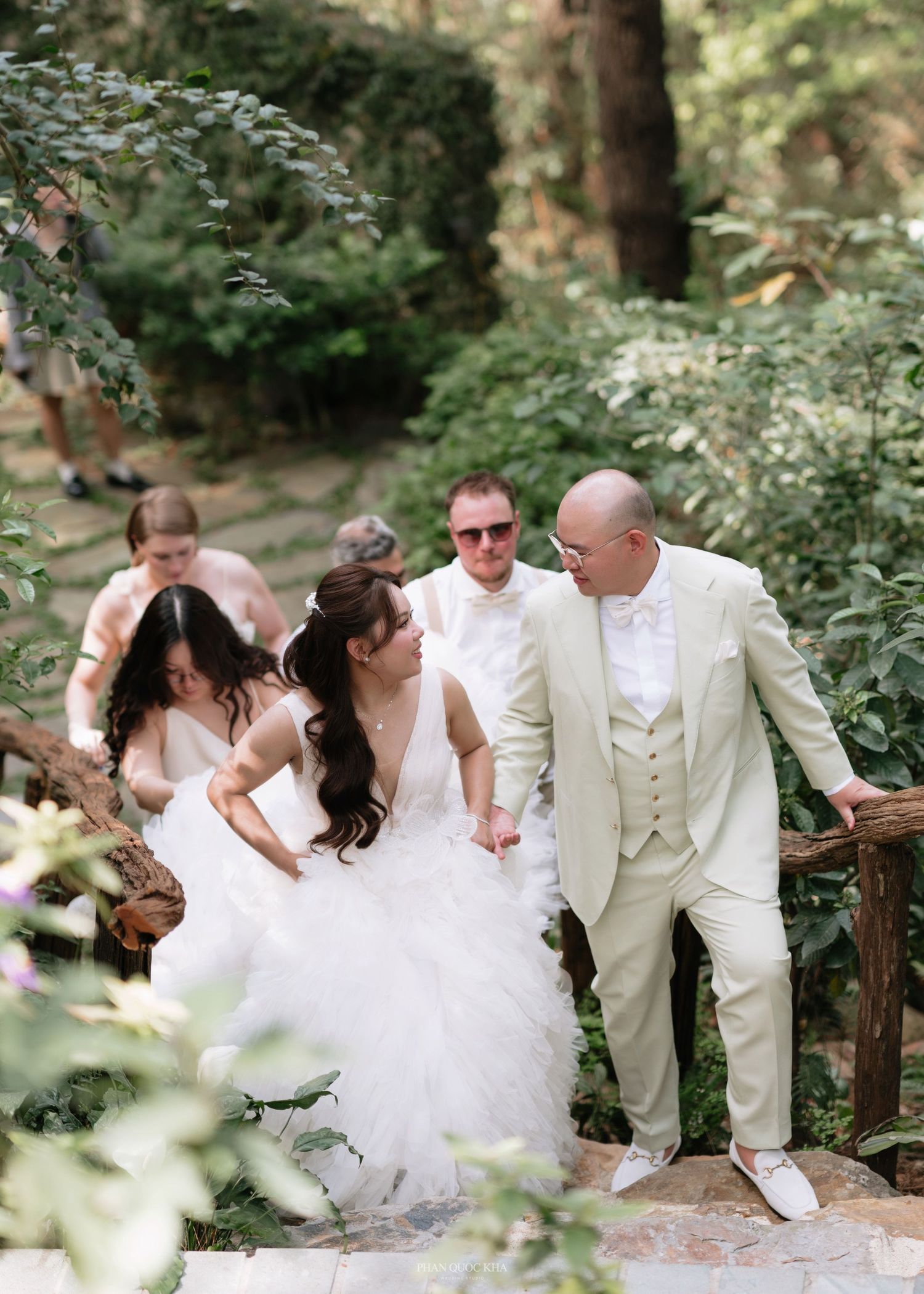 Bride and groom walking down stone steps in a lush green garden with wedding party.
