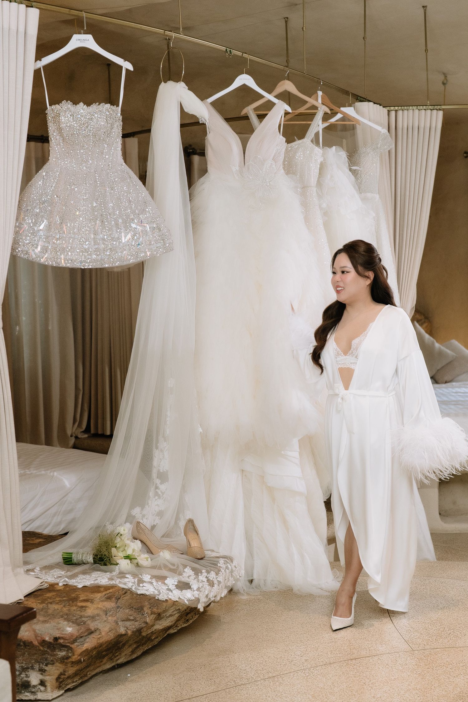 Bride in white feather robe admires row of elegant wedding dresses hanging in a bridal boutique.