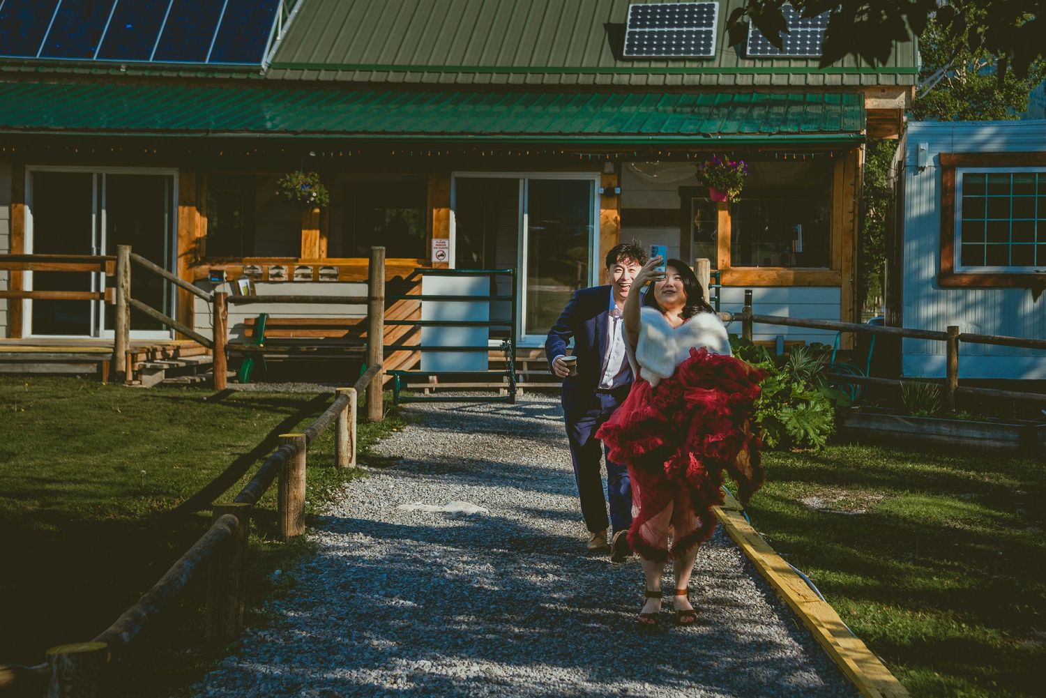 Two people stroll down a gravel path leading to a green-roofed rustic building on a sunny day. Alberta Helicopter Elopement Photographer