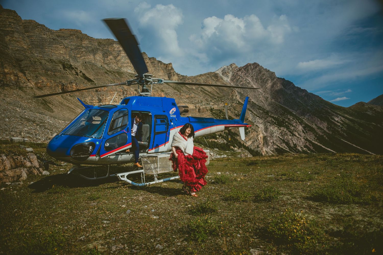 Helicopter parked in a mountainous valley with rocky cliffs and green grass. Alberta Helicopter Elopement Photographer