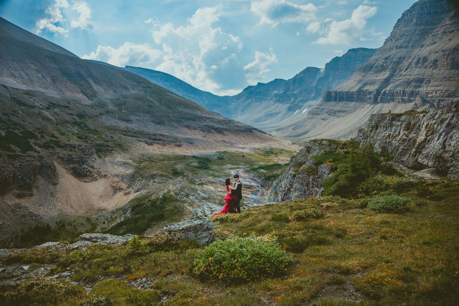 A figure in red stands on a rocky mountain overlook with dramatic cliffs and valleys Alberta Helicopter Elopement Photographer