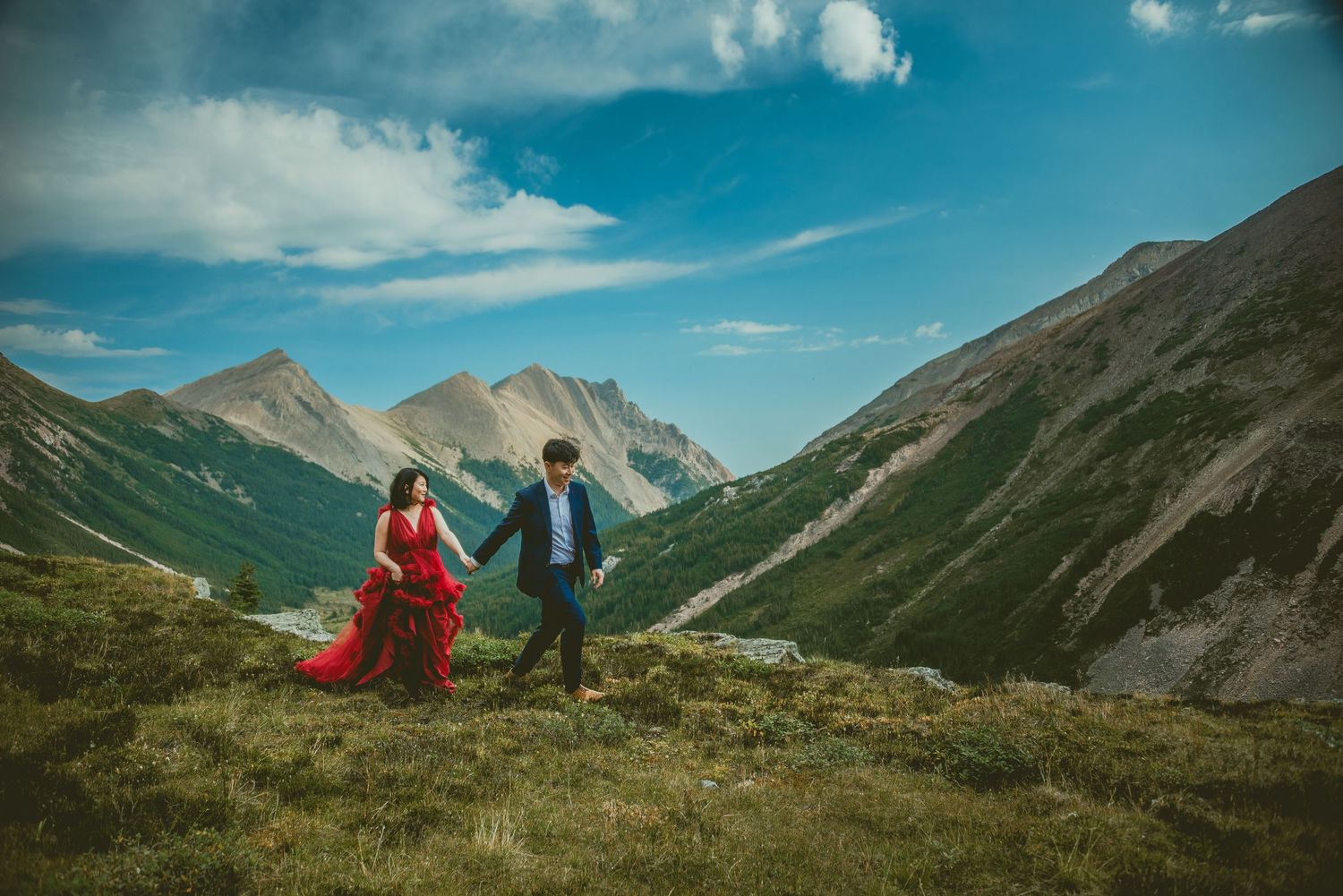 A couple stands atop a grassy mountain ridge with dramatic peaks and blue skies in the background. Alberta Helicopter Elopement Photographer