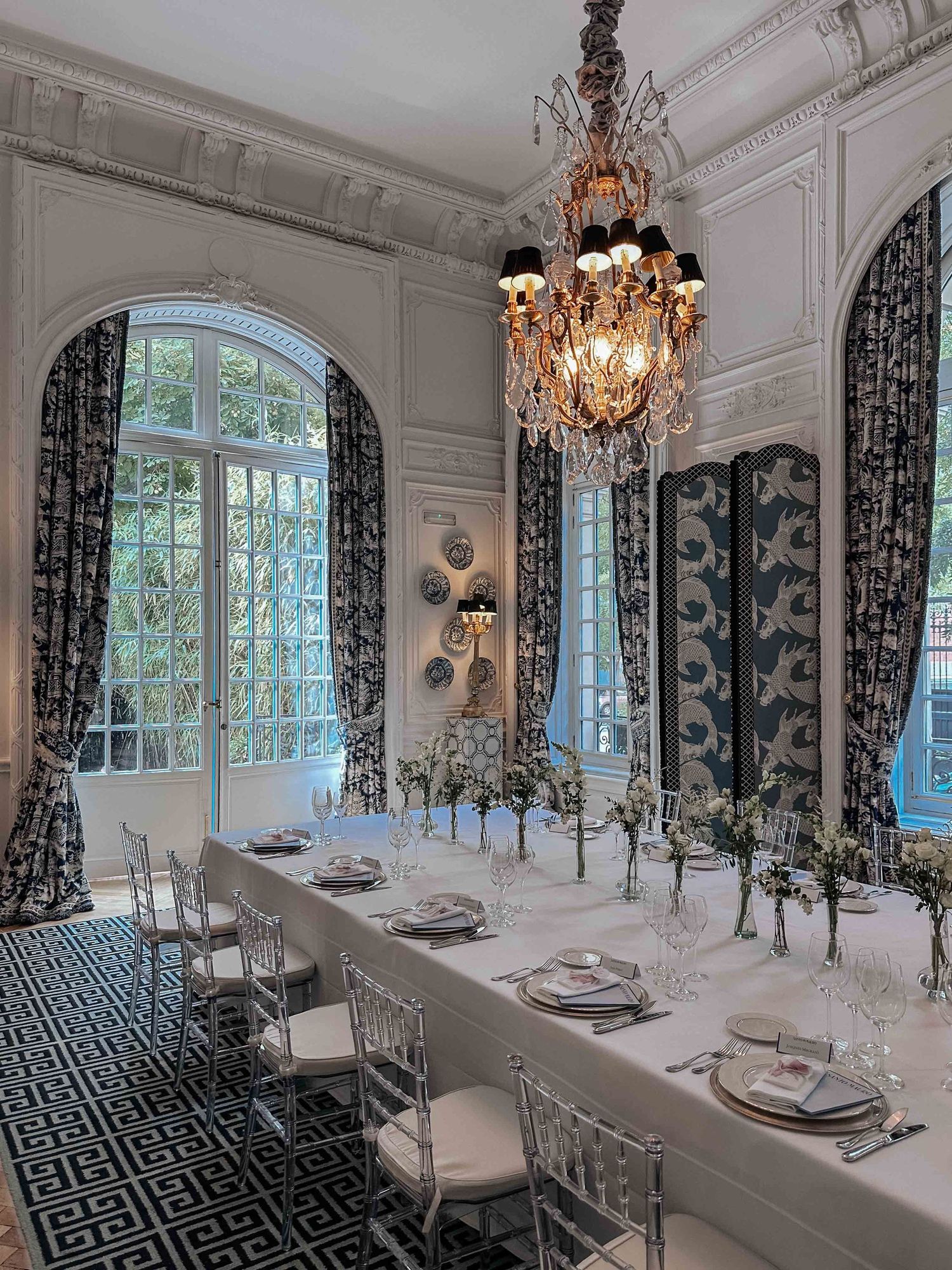 Elegant dining room with crystal chandeliers, arched windows, patterned carpet and luxurious table settings in white and gold.