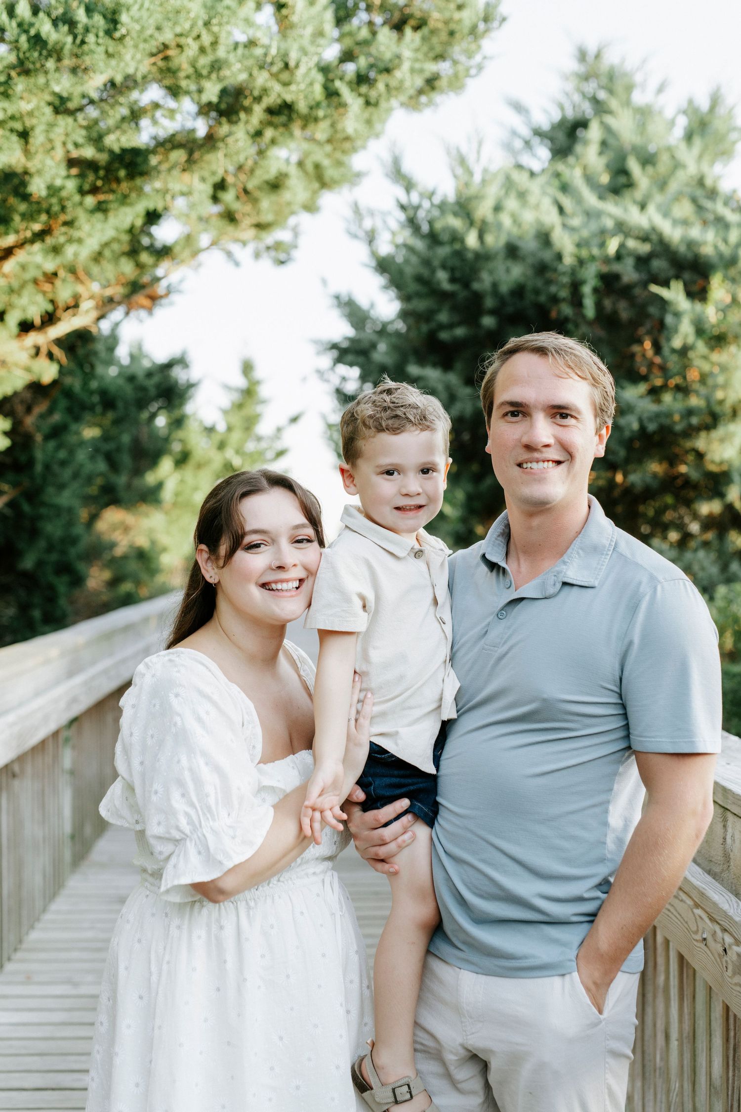 Family poses together on a wooden boardwalk surrounded by evergreen trees on a bright sunny day.