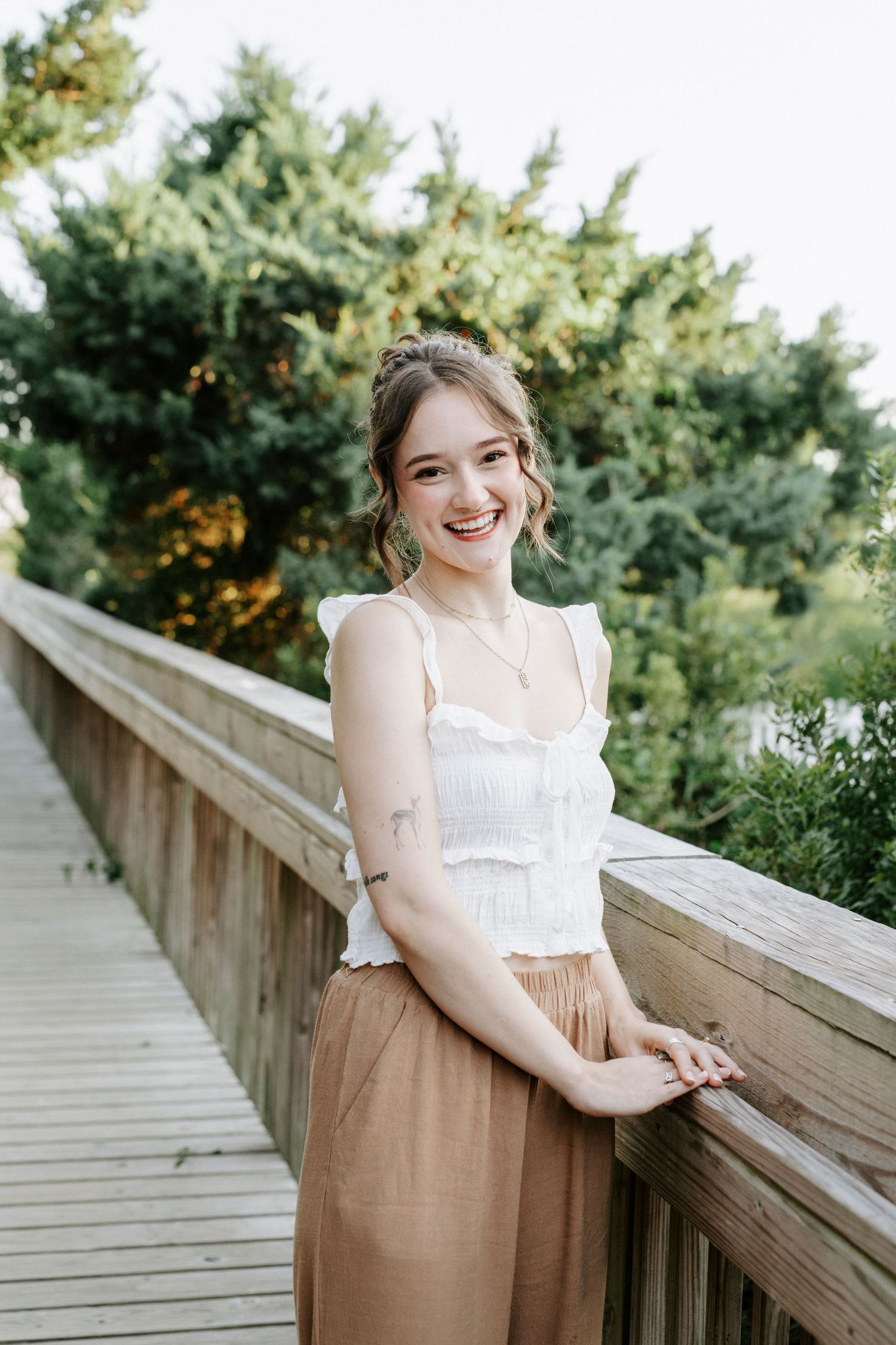 Young person in white crop top and tan skirt smiling and posing along a wooden railing at an outdoor location.