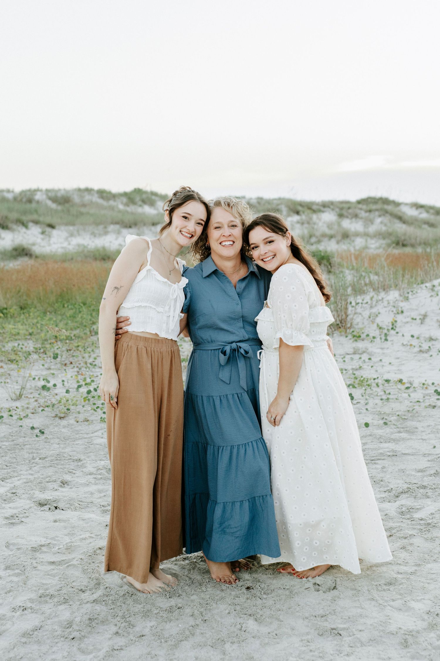 Three friends in flowing dresses embrace and smile together on a sandy beach at sunset.