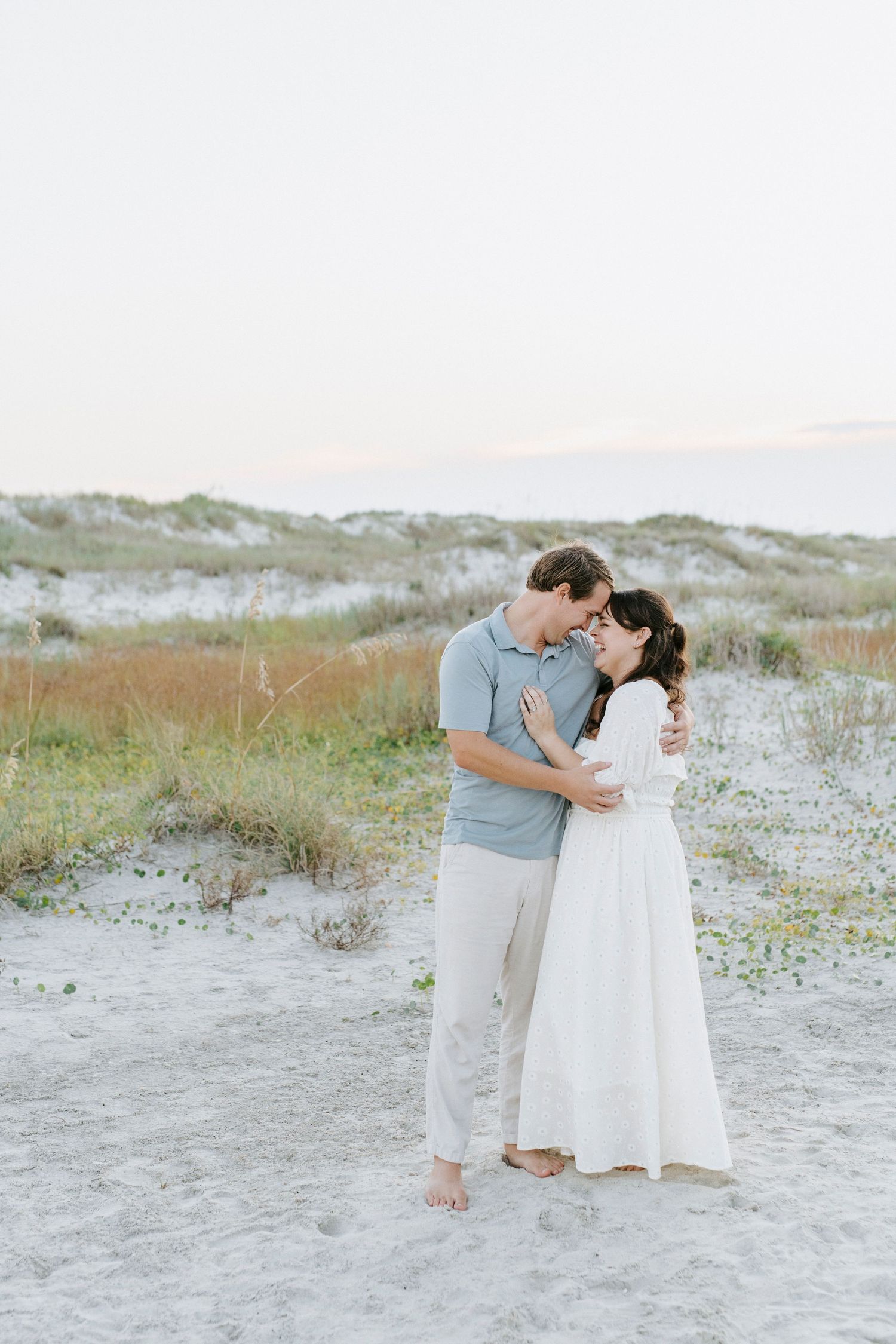 A romantic coastal engagement photo of a couple embracing on a sandy beach with dunes and grass in the background.