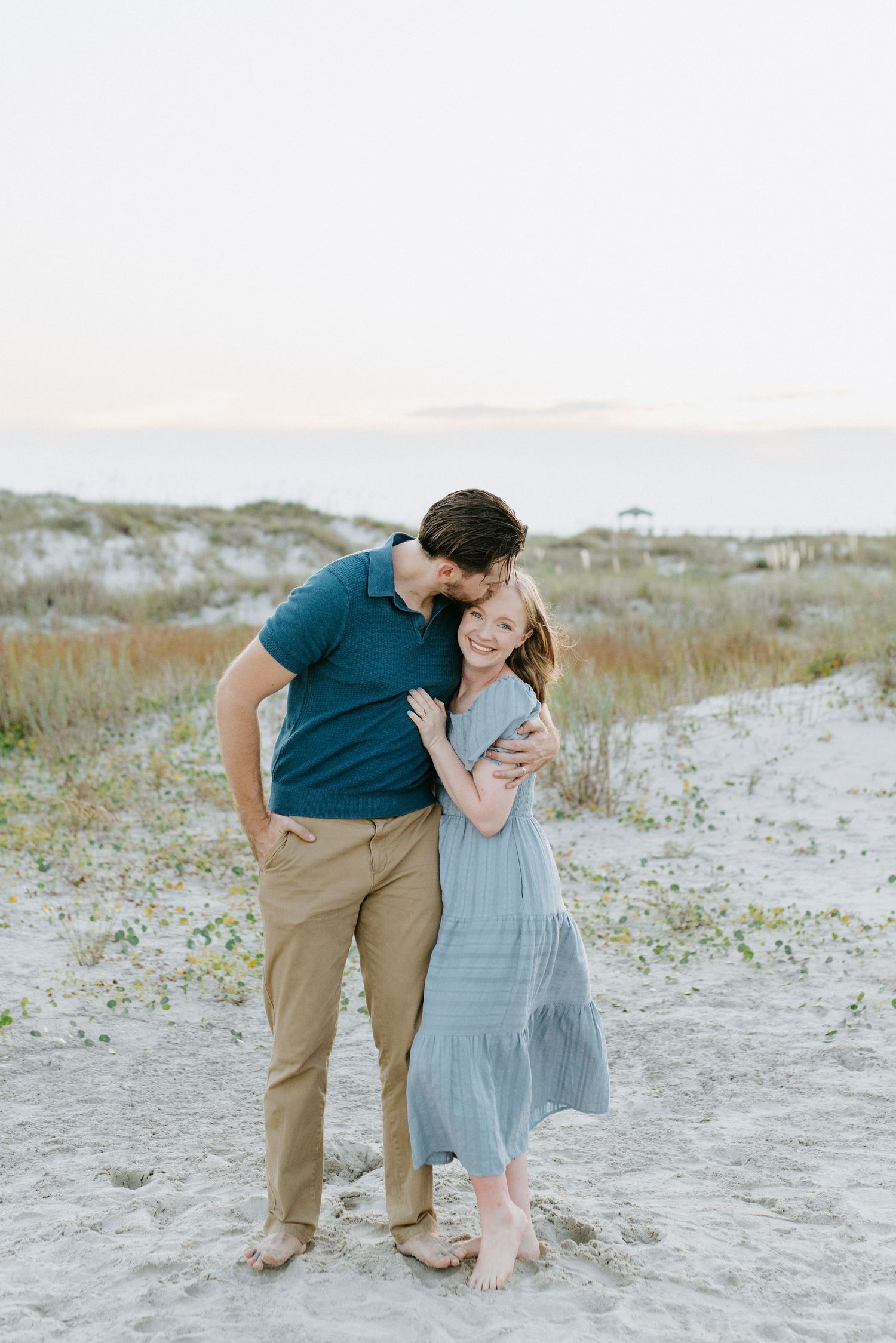 A romantic couple embracing on a sandy beach with dunes in the background.
