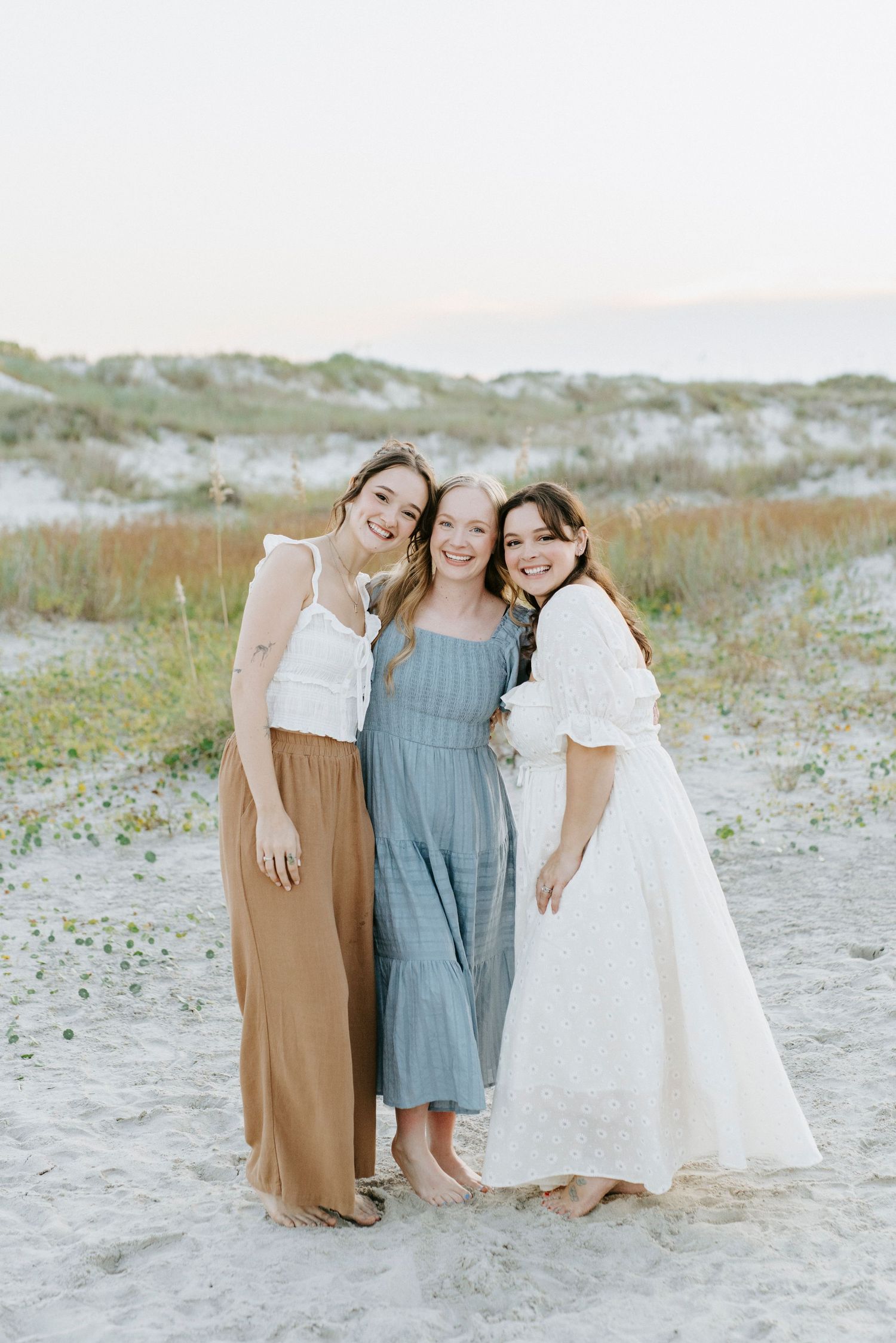 Three friends wearing coordinated dresses embrace and laugh together on a sandy beach at sunset.
