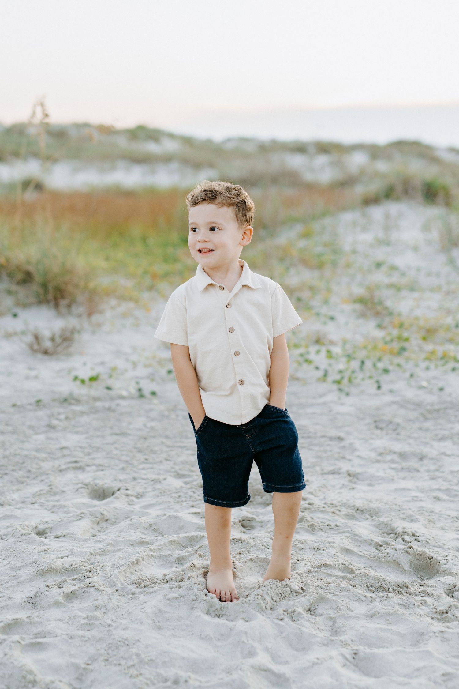 A young child poses on sandy beach wearing a light colored shirt and navy shorts.