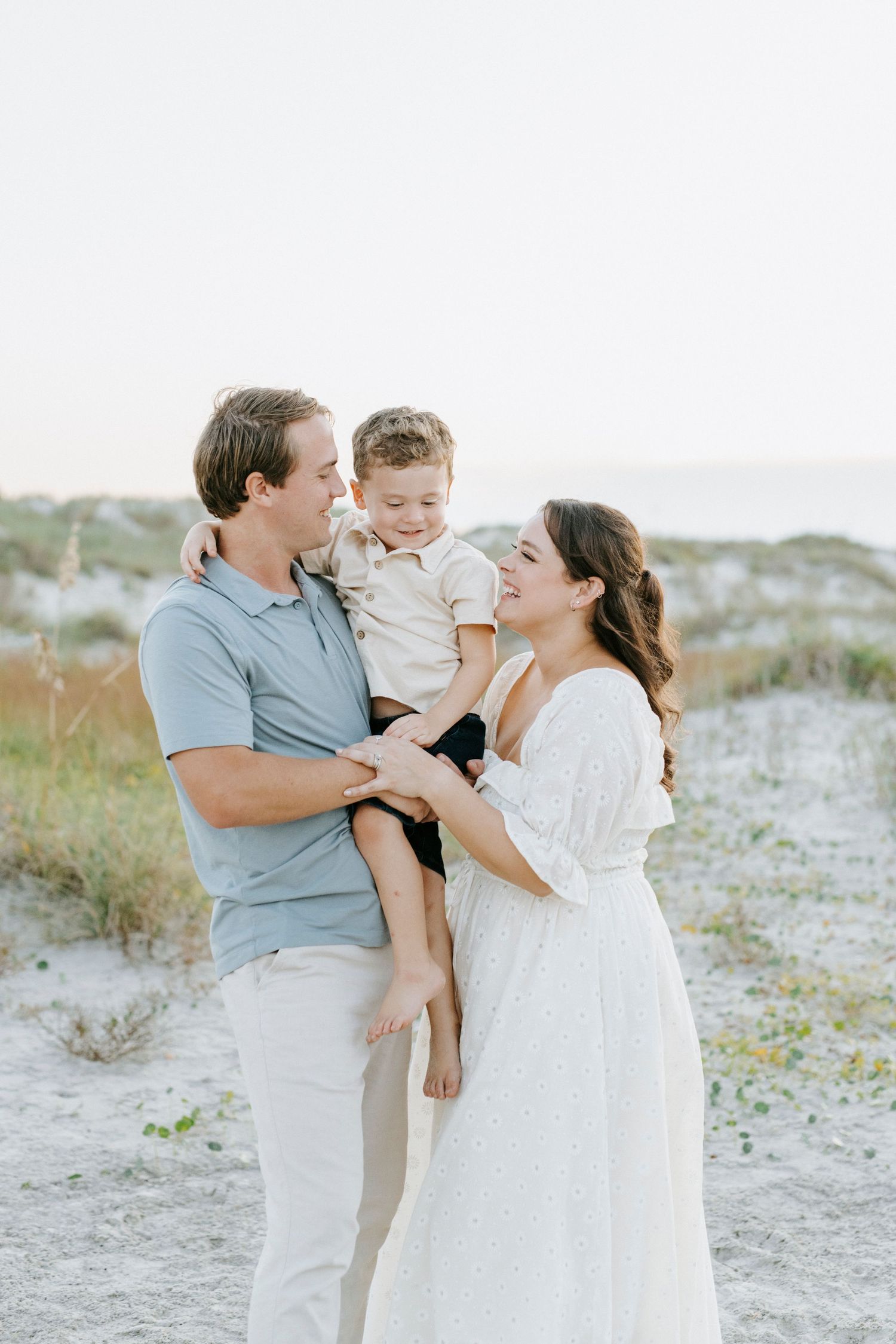 Family shares tender moments together on a breezy beach with coastal vegetation in the background.
