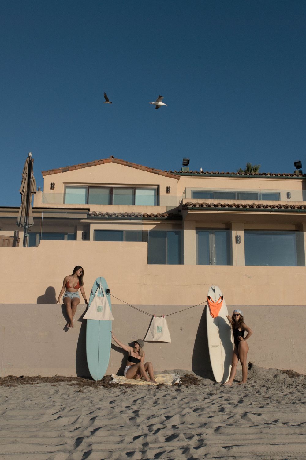 Surfers with surfboards relaxing on a sandy beach at sunset near coastal buildings.