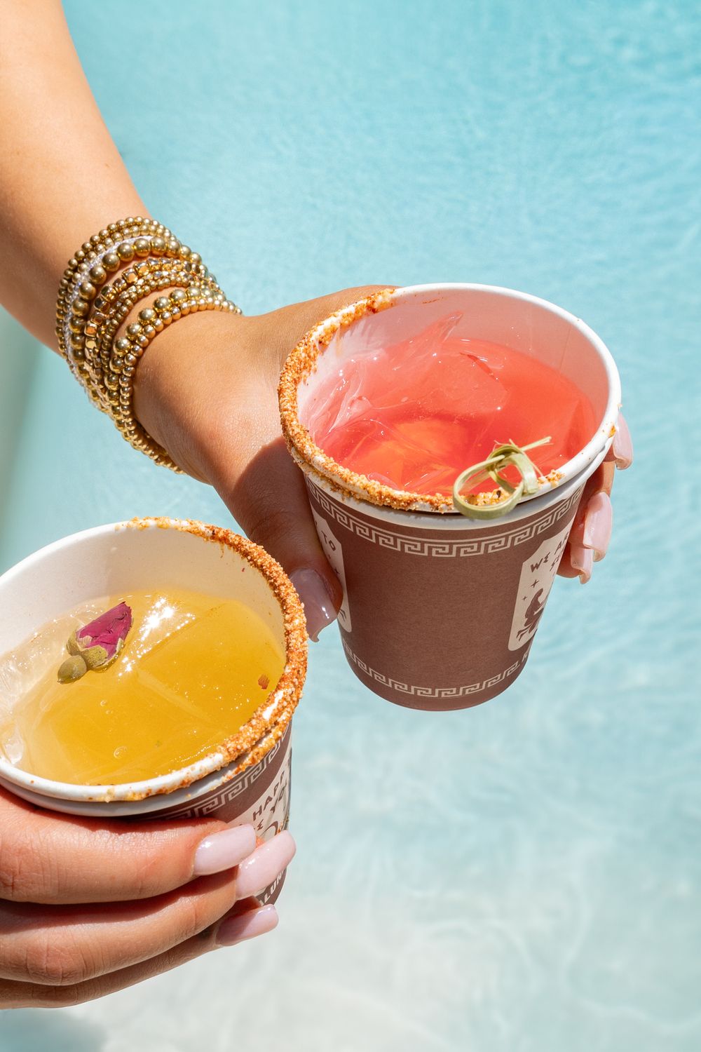Colorful cocktails in decorative glasses held by hands adorned with gold bangles against turquoise background.