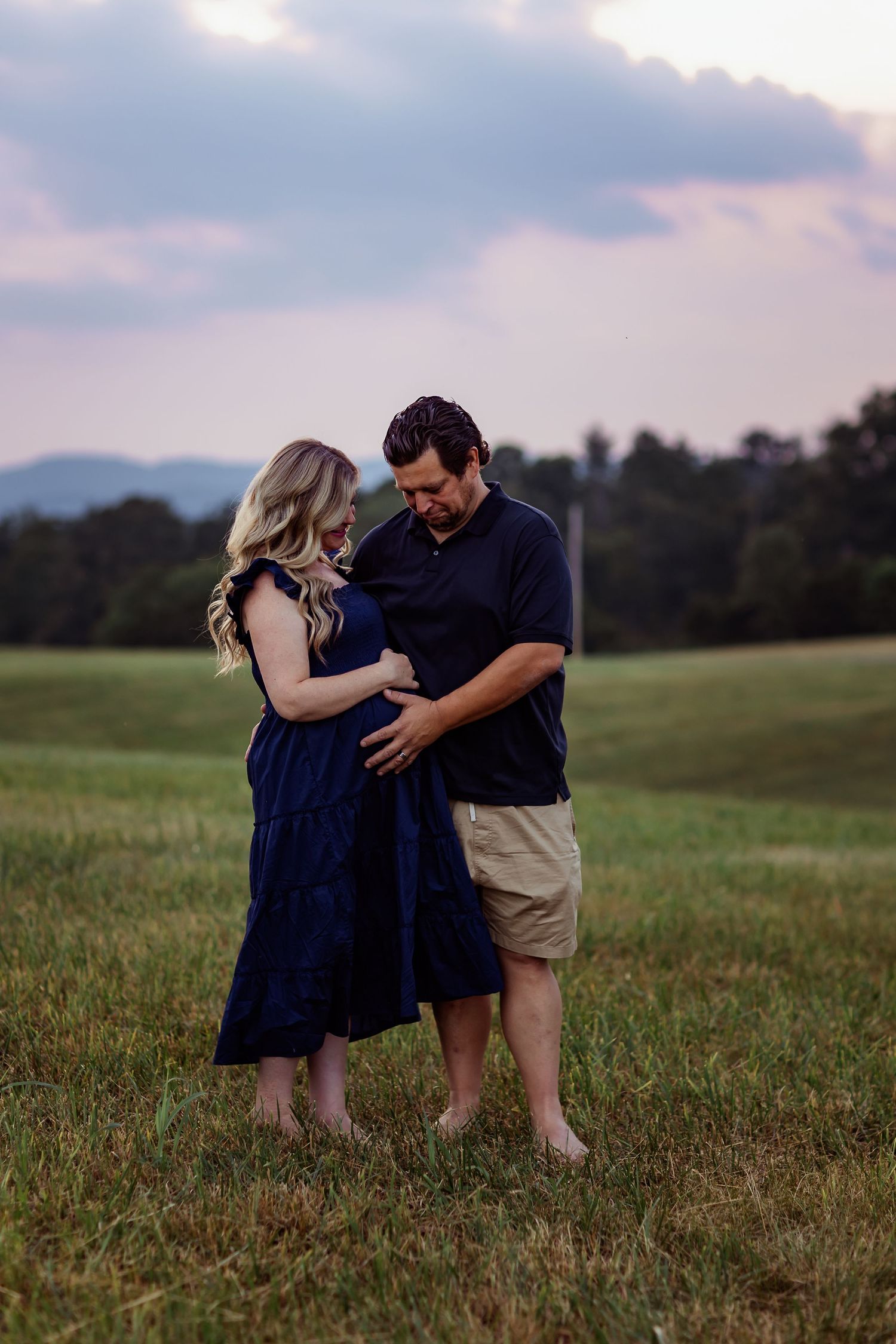 A couple shares an intimate moment together in a grassy field at dusk with mountains in the background.