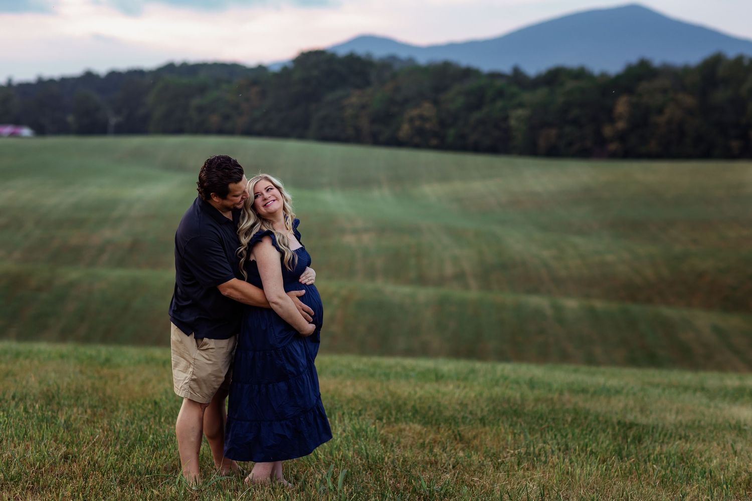 Two people share an intimate moment in a scenic green meadow with mountains in the background.