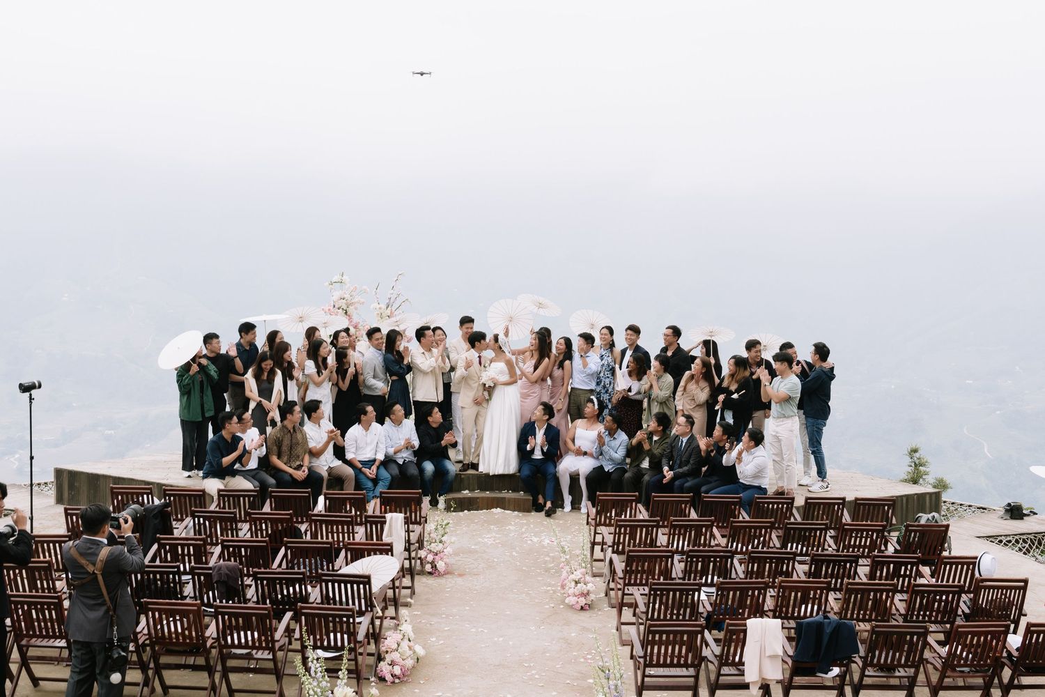 Wedding party group photo at scenic mountain ceremony venue with rows of wooden chairs and floral decor.