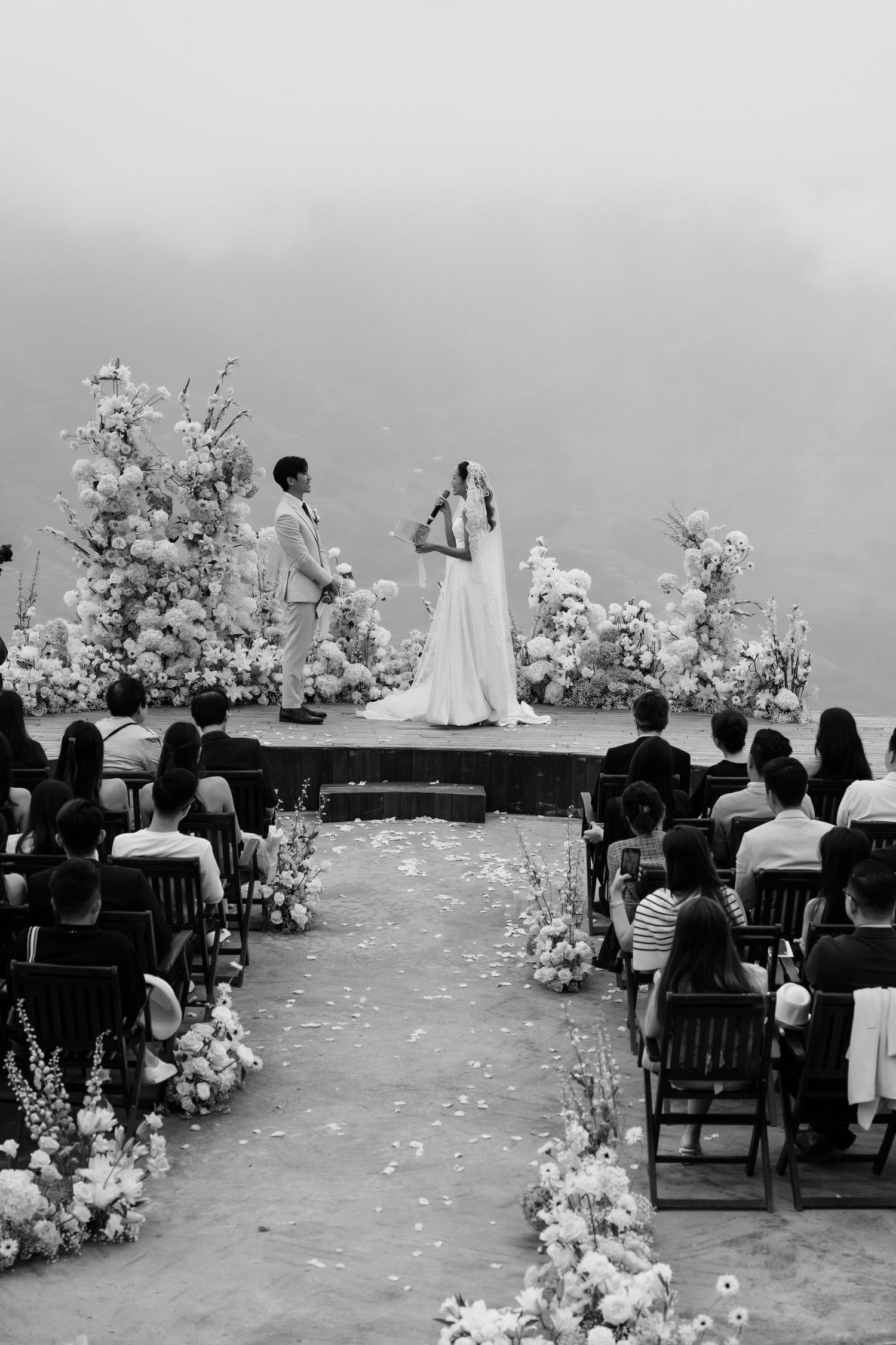 Black and white wide shot of a couple exchanging vows at a floral outdoor ceremony with seated guests on a misty day.