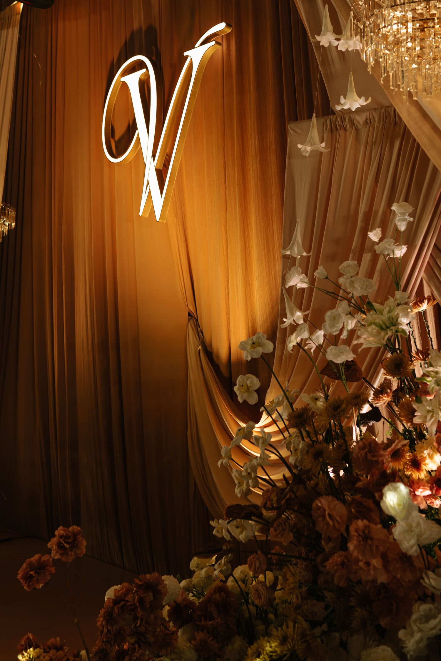 Elegant wedding venue backdrop with illuminated monogram letter V, draped golden curtains, and floral arrangements.