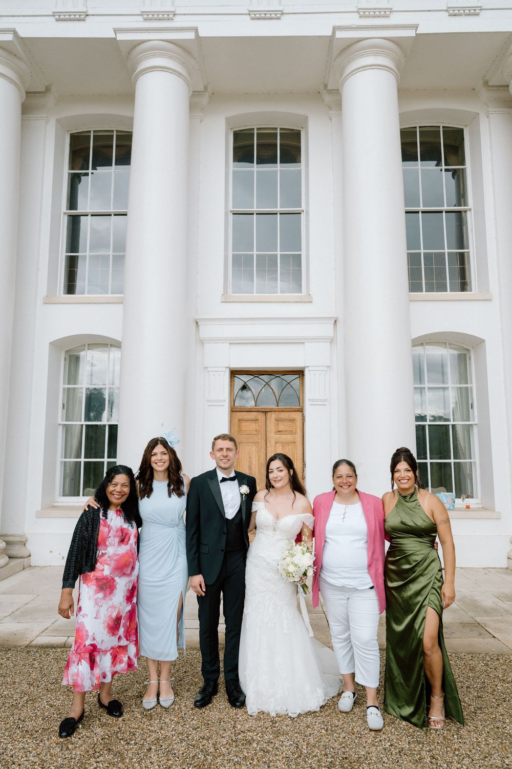 Wedding party poses together in formal attire against white pillared building exterior. Taken by modern editorial wedding photographer.
